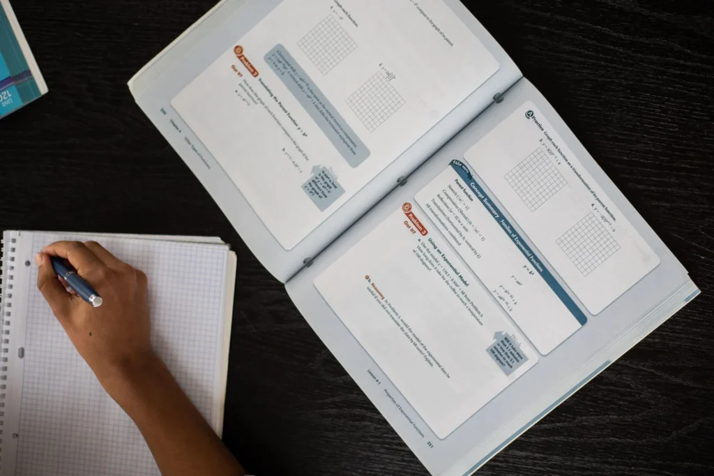 An open math textbook and notebook with a hand writing