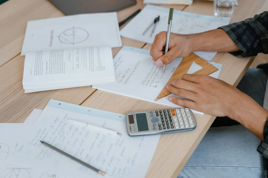 A person writing math notes next to an open textbook and calculator