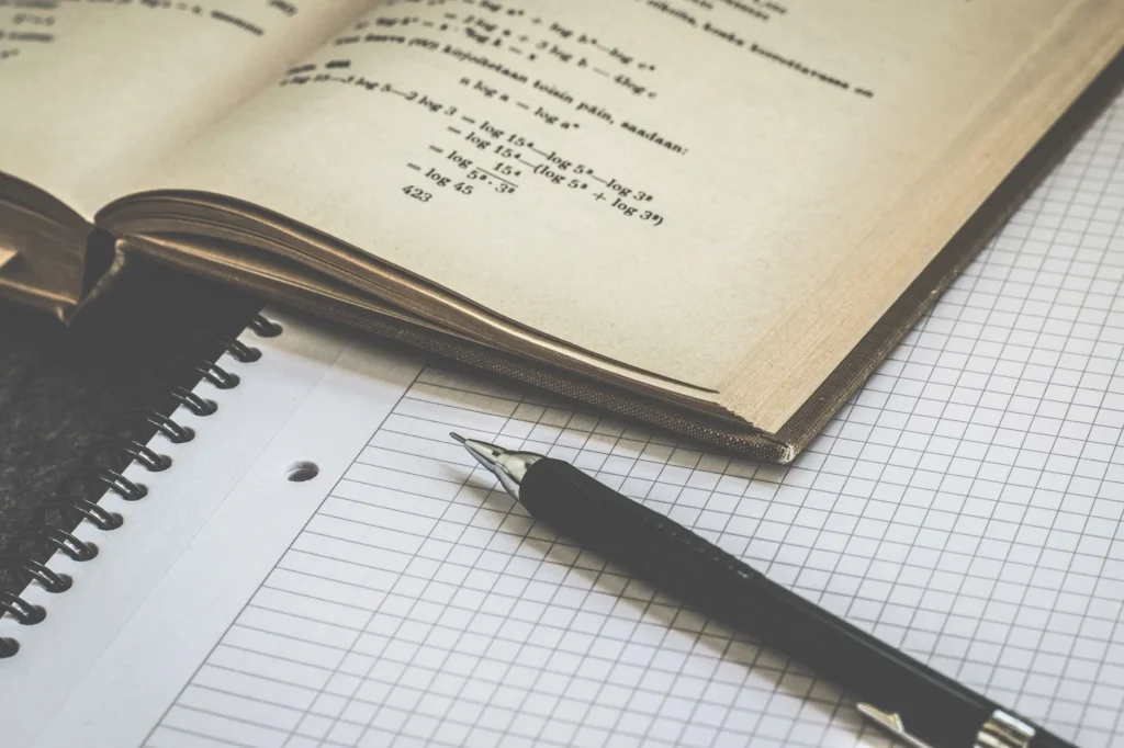 An open textbook, a notebook, and a pen on a grid paper.