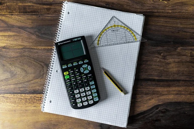 A graphing calculator, protractor, pencil, and grid notebook on wooden table