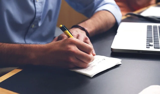 A person writing notes with a pen next to a laptop.